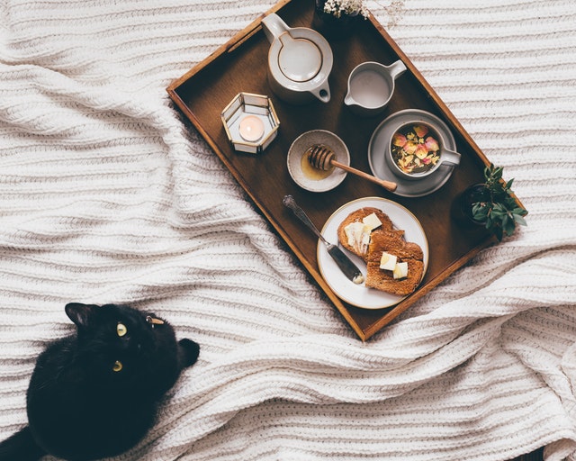 A black cat sitting in front of a tray with a breakfast on a bed.