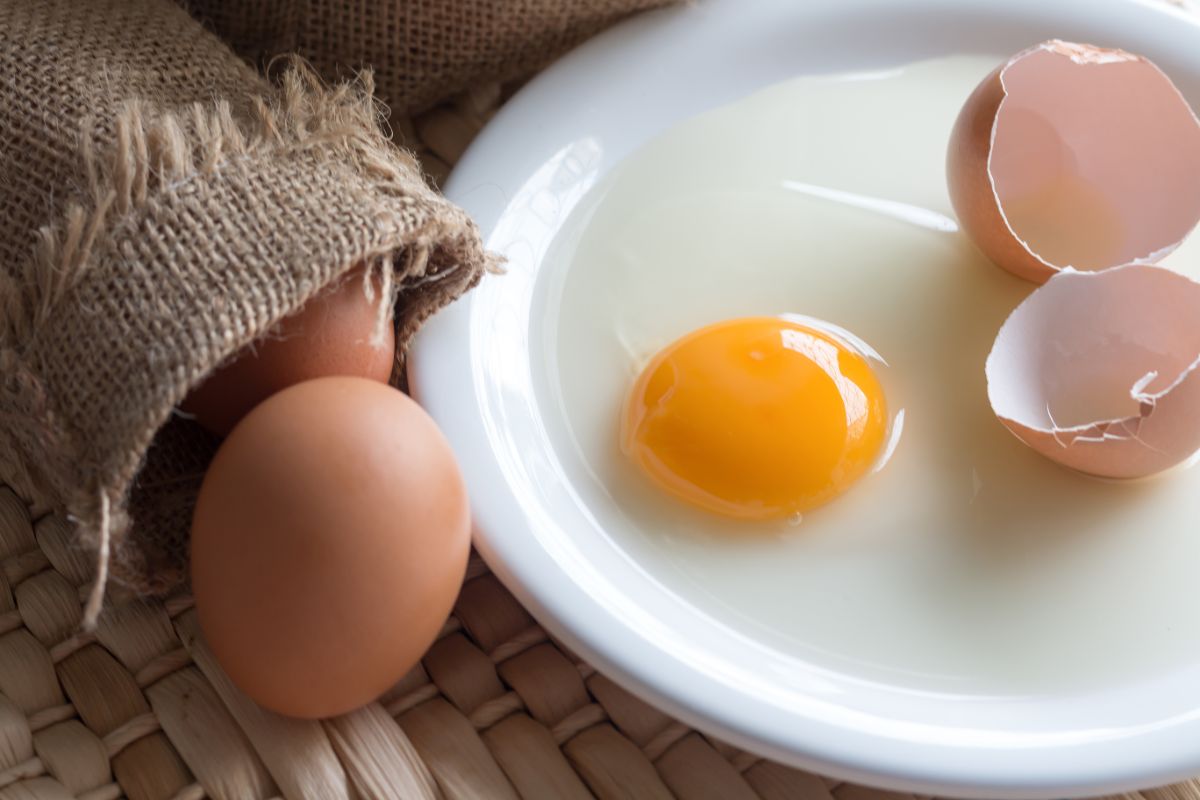 A broekn egg with shells on a whie plate next to a sack of whole eggs.