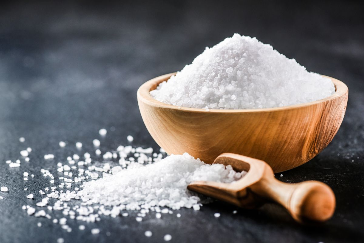 A wooden bowl full of salt and a wooden scoop with saltk on a black table.