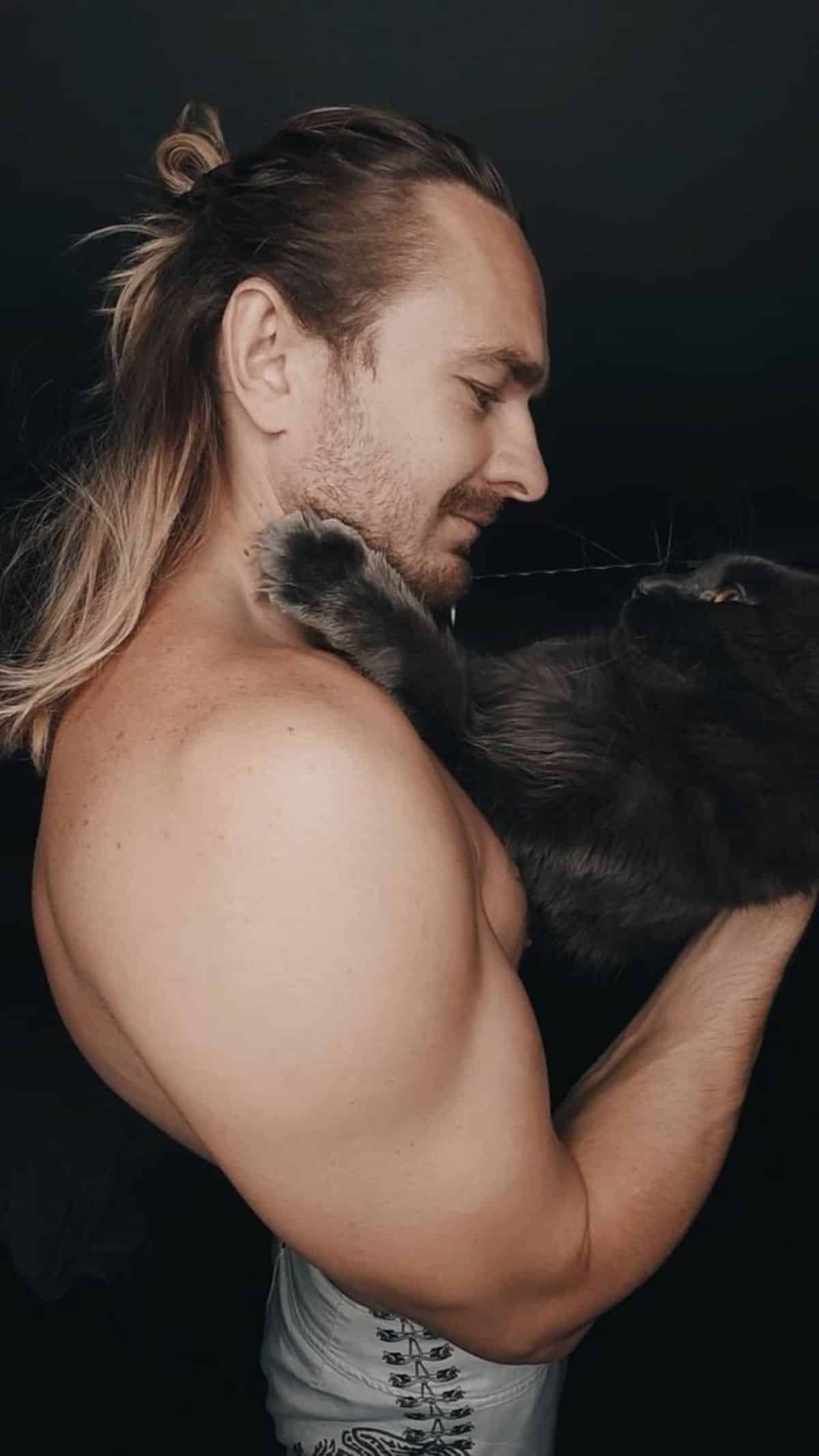 A topless man holding a huge black maine coon.