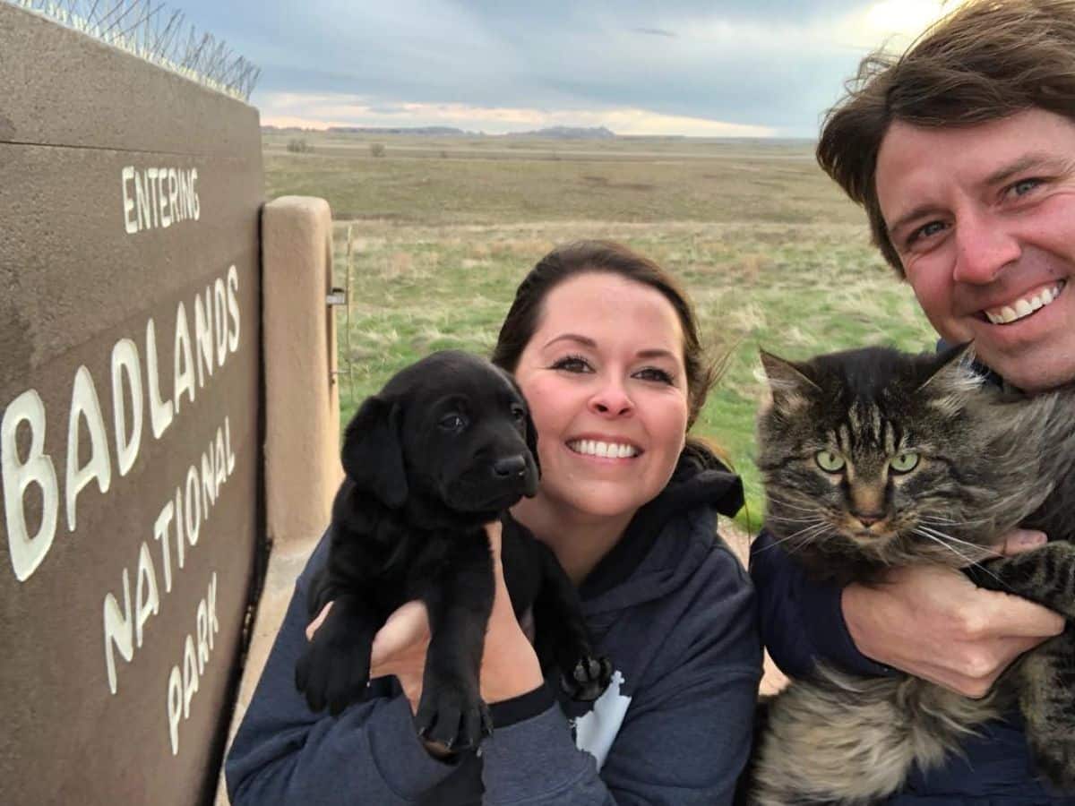 A married couple holding a black labrador puppy and a tabby maine coon nead a national park sign.